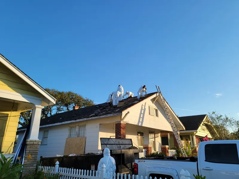 Roofing crew in protective coveralls working on a roof tear-off beside a ladder and debris container